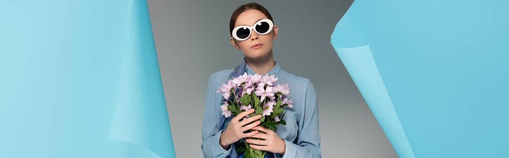 glamour woman in sunglasses holding pink flowers near hole in blue paper on grey background, banner - Photo, Image