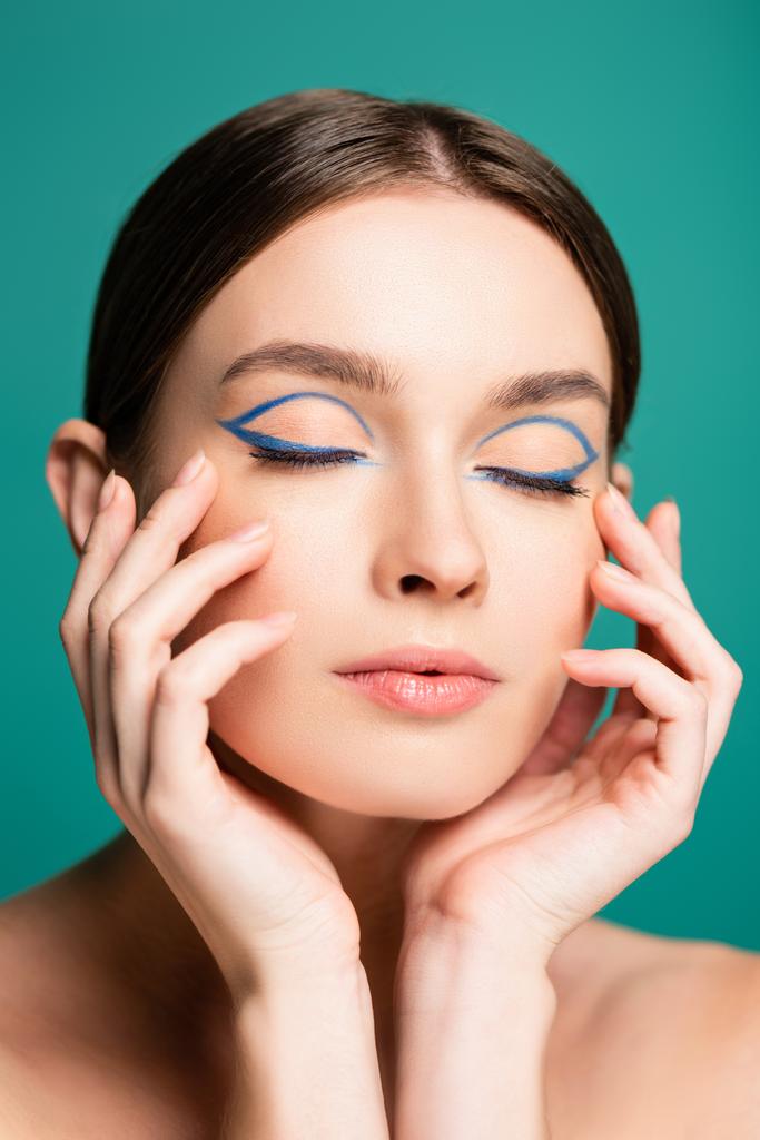 urocza kobieta z niebieskim eyeliner w zamkniętych oczach pozowanie z dłońmi blisko twarzy odizolowane na zielono - Zdjęcie, obraz