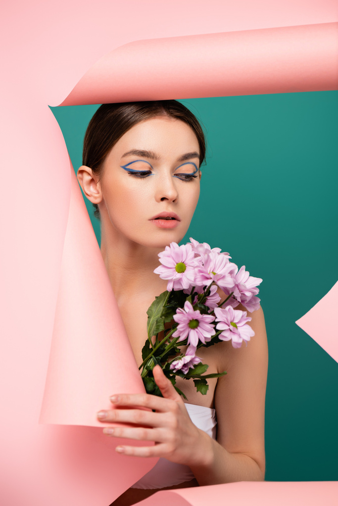 hezká žena s kreativním make-up držení chryzantémy v blízkosti otvoru v růžovém papíru izolované na zelené - Fotografie, Obrázek