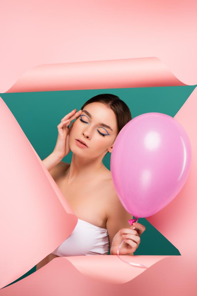 young woman with closed eyes holding balloon near hole in pink paper isolated on green - Photo, Image