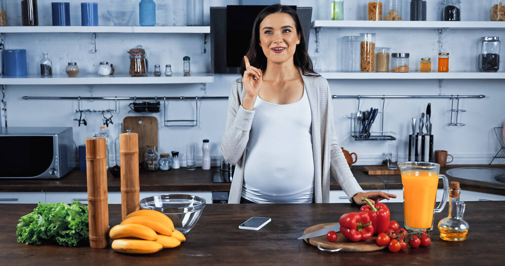 Donna incinta che ha un'idea vicino smartphone e cibo fresco sul tavolo della cucina - Foto, immagini