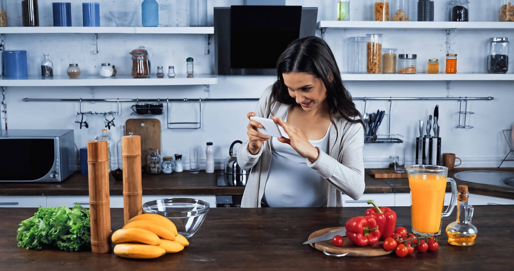 Femme enceinte souriante prenant des photos avec smartphone près d'ingrédients frais sur la table de cuisine  - Photo, image