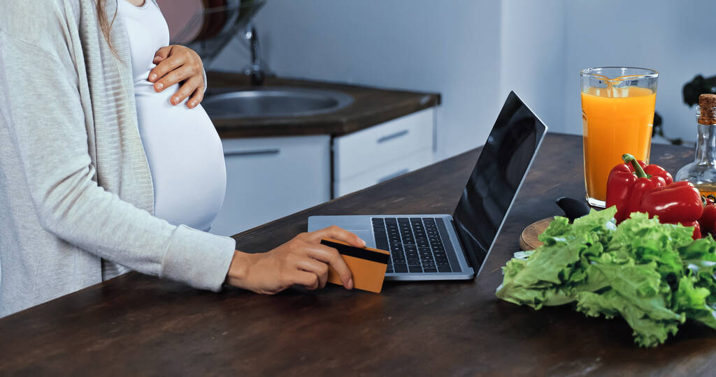 Vue recadrée d'une femme enceinte tenant une carte de crédit près d'un ordinateur portable et de légumes  - Photo, image