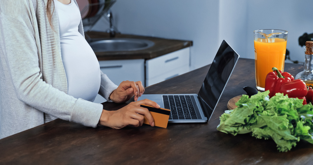 Vista ritagliata di donna incinta in possesso di carta di credito vicino al computer portatile e cibo fresco in cucina  - Foto, immagini
