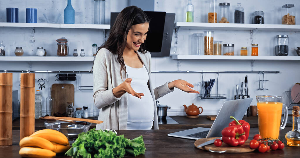 Vidám terhes nő, amelynek videó hívás a laptopon, közel élelmiszer a konyhában  - Fotó, kép