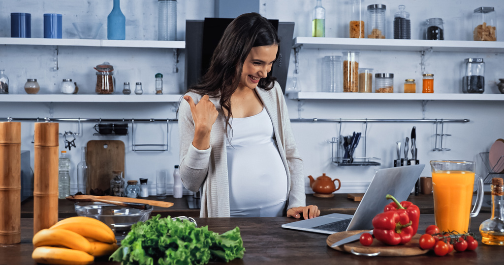 Χαμογελώντας έγκυος γυναίκα δείχνει σαν κατά τη διάρκεια βιντεοκλήσης κοντά λαχανικά στην κουζίνα  - Φωτογραφία, εικόνα