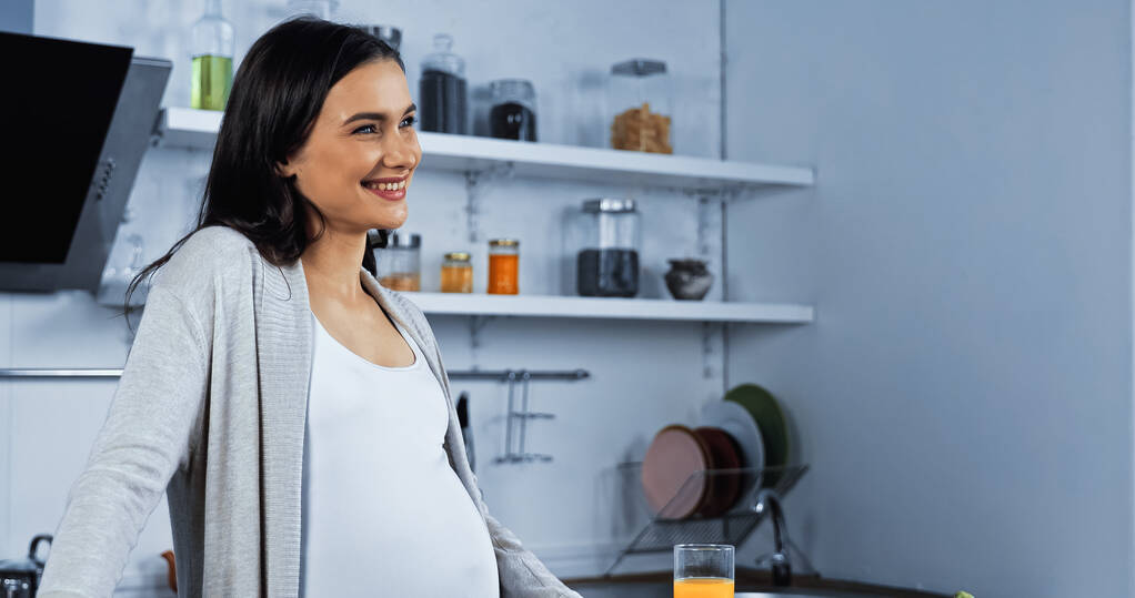 Mujer embarazada sonriente mirando lejos cerca del jugo de naranja en la cocina  - Foto, imagen