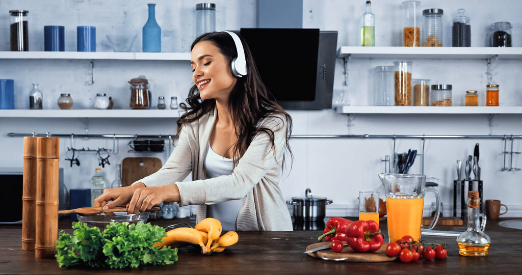 Mosolygó nő fülhallgatóban áll friss étel mellett a konyhában  - Fotó, kép