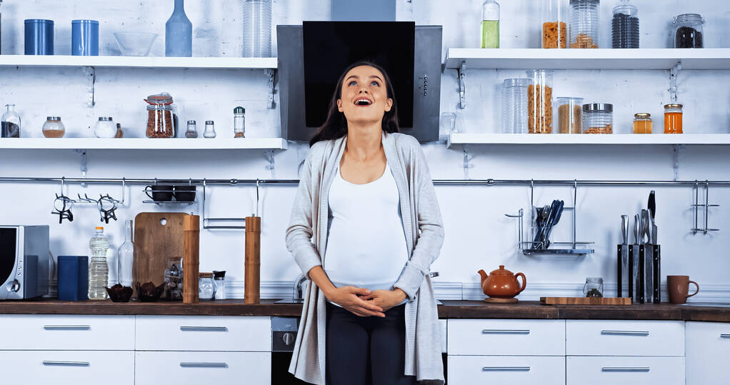 opgewonden zwangere vrouw omarmen buik in keuken  - Foto, afbeelding
