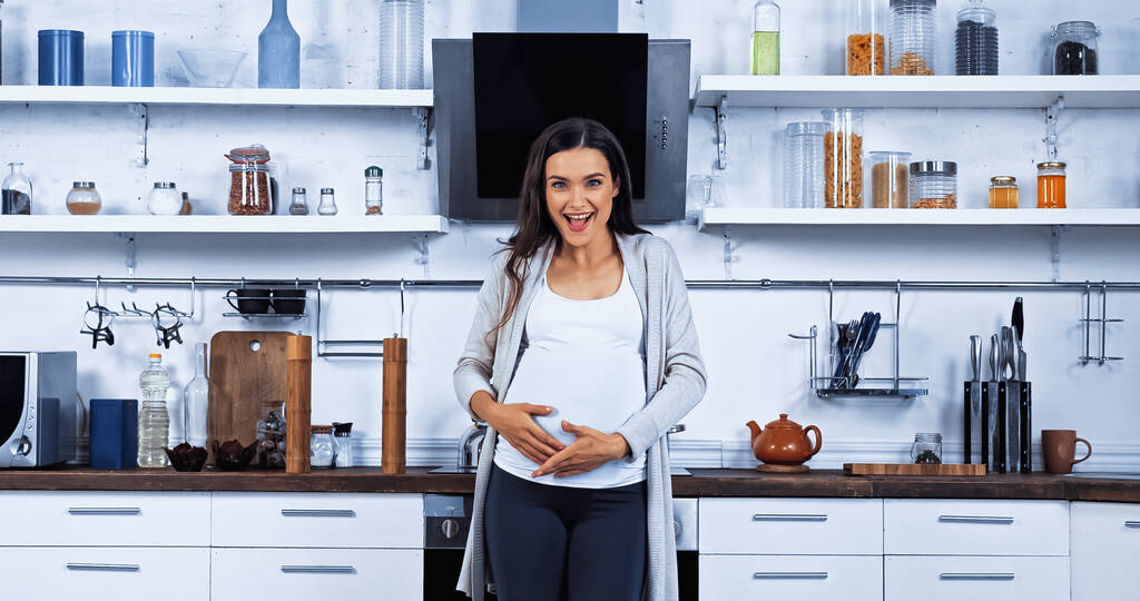 Iloinen raskaana oleva nainen katsoo kameraa keittiössä  - Valokuva, kuva