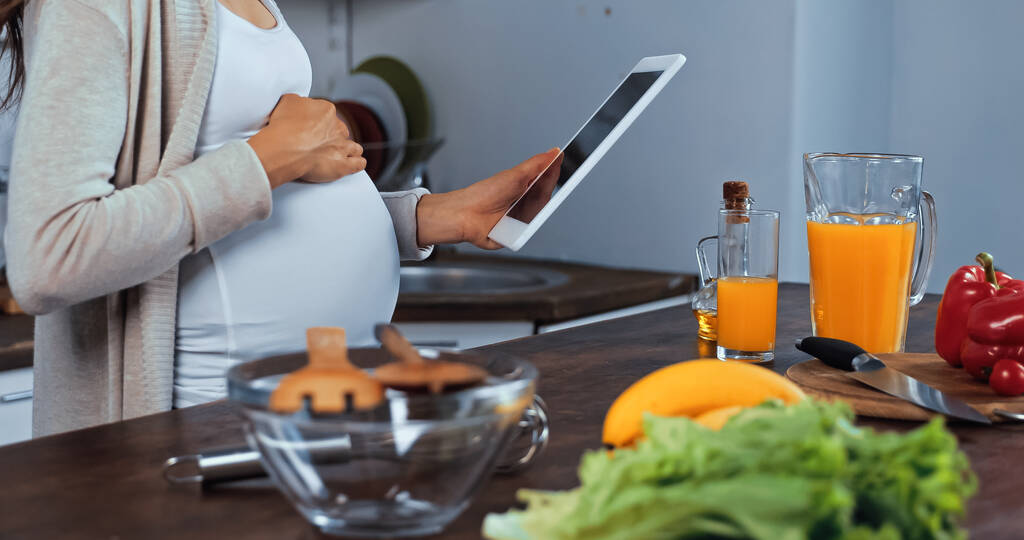 Ausgeschnittene Ansicht einer schwangeren Frau mit digitalem Tablet in der Nähe von Gemüse und Orangensaft in der Küche  - Foto, Bild