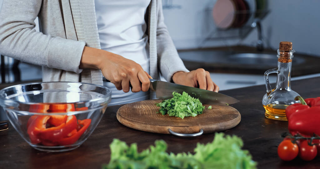 Обрізаний вид на жінку, що ріже листя салату біля миски та овочів на розмитому фоні
  - Фото, зображення