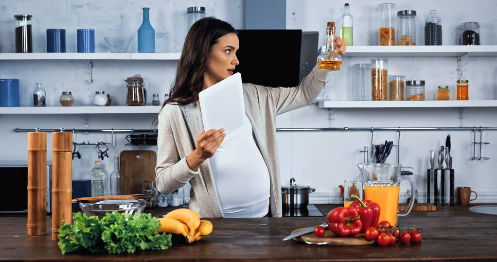 Schwangere mit digitalem Tablet schaut Öl in der Nähe von Lebensmitteln auf Küchentisch  - Foto, Bild