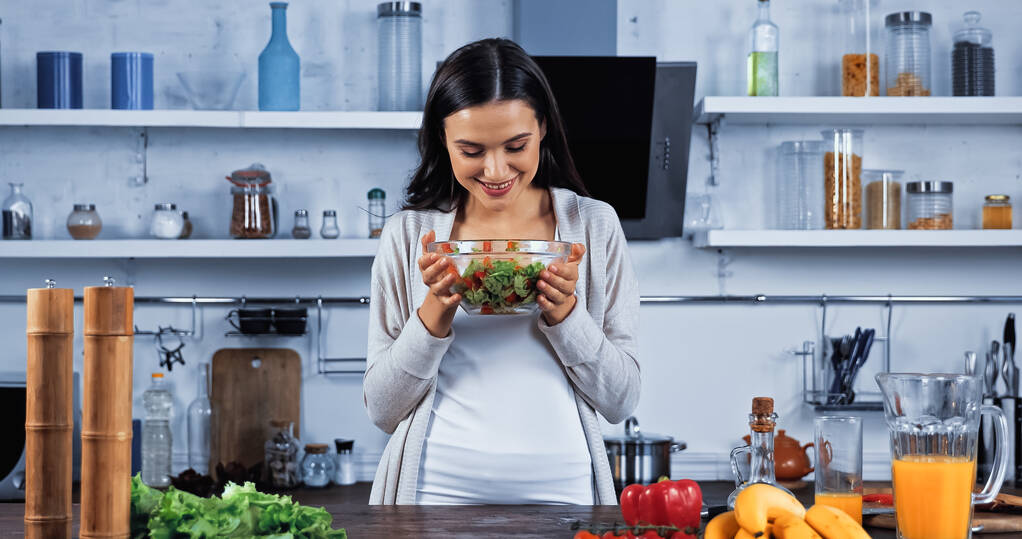 Mujer sonriente mirando ensalada fresca cerca de ingredientes orgánicos y jugo de naranja  - Foto, Imagen