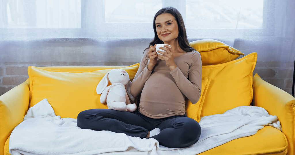 Mulher grávida sonhadora com copo sentado perto de brinquedo macio  - Foto, Imagem
