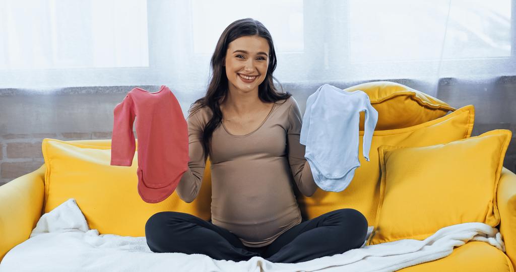 Mujer embarazada sonriente sosteniendo ropa de bebé y mirando a la cámara  - Foto, Imagen