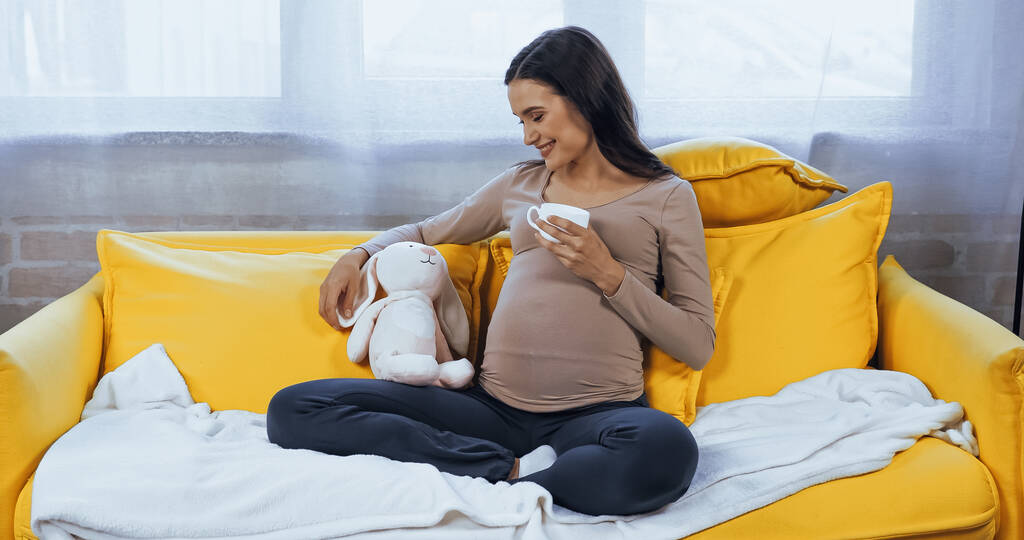 妊娠中女性とともにカップ見ます柔らかいおもちゃに座っている間に黄色のソファ  - 写真・画像