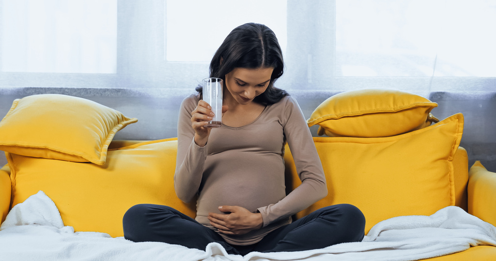 Donna incinta che tiene un bicchiere di latte fresco e guarda la pancia sul divano  - Foto, immagini