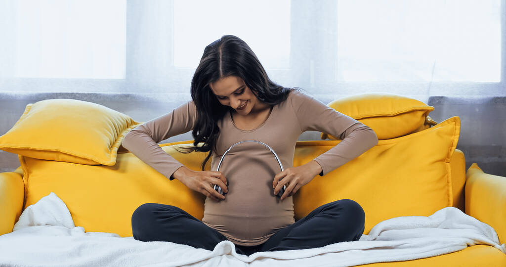 Happy pregnant woman holding headphones near belly while sitting on couch  - Photo, Image