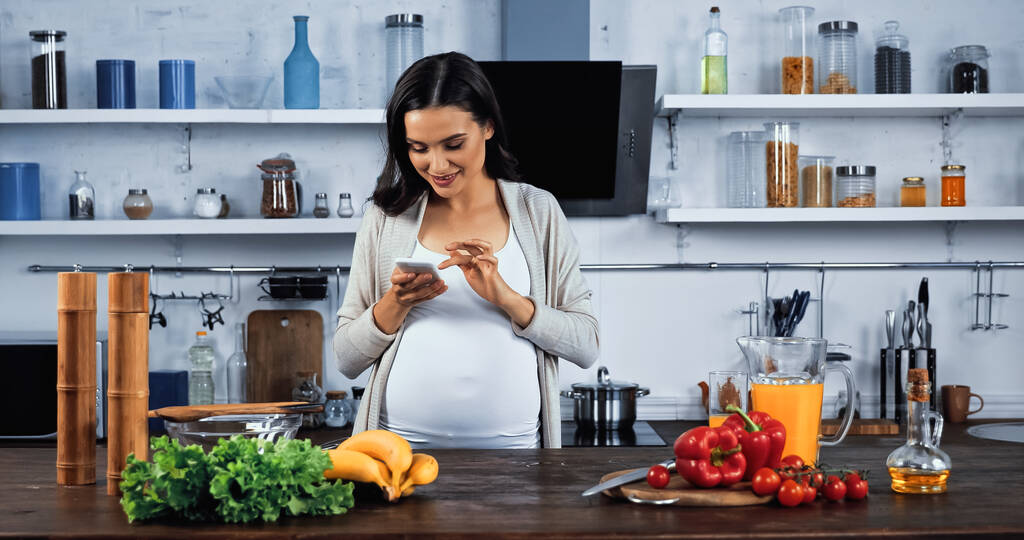 Smiling pregnant woman using smartphone near raw food on kitchen table  - Photo, Image