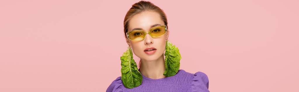 young woman in colored eyeglasses and fresh lettuce earring isolated on pink, banner - Photo, Image