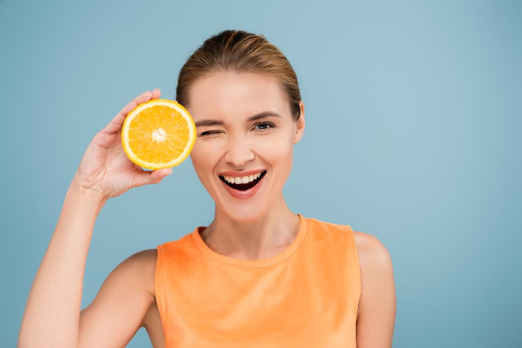 excited woman winking at camera while holding half of ripe orange isolated on blue - Photo, Image