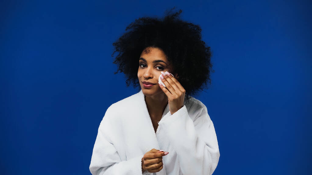 Femme afro-américaine nettoyage visage avec tampon de coton isolé sur bleu  - Photo, image