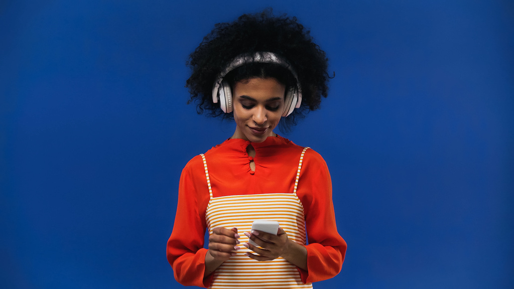 Αφροαμερικανή γυναίκα που χρησιμοποιεί ακουστικά και smartphone απομονώνονται σε μπλε - Φωτογραφία, εικόνα