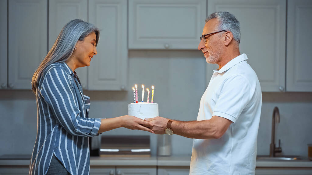 vue latérale de sourire senior homme présentant gâteau d'anniversaire à joyeuse asiatique femme - Photo, image