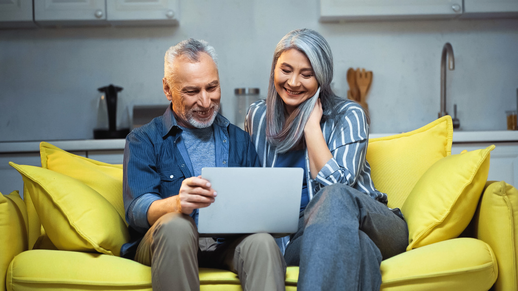 heureux couple interracial assis sur canapé avec ordinateur portable à la maison - Photo, image