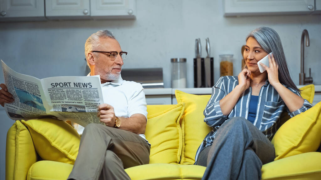 пожилой мужчина с газетой смотрит на азиатскую жену разговаривающую на смартфоне - Фото, изображение