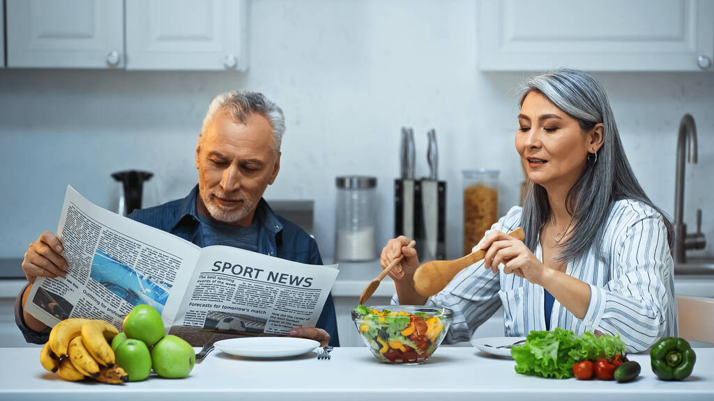 пожилая азиатка, смешивающая салат с мужем, читает спортивные новости во время завтрака - Фото, изображение