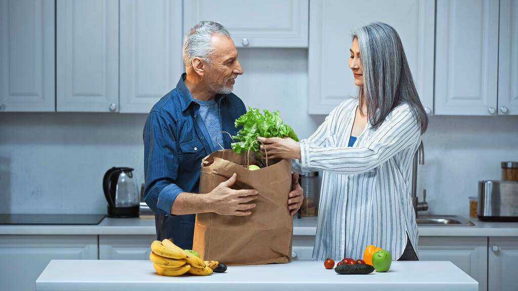 пожилая межрасовая пара распаковывает бумажный пакет со свежими овощами и фруктами на кухне - Фото, изображение