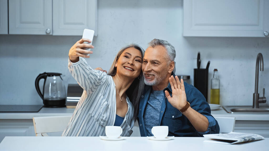 veselý starší muž mává rukou v blízkosti asijské manželka přičemž selfie v kuchyni - Fotografie, Obrázek