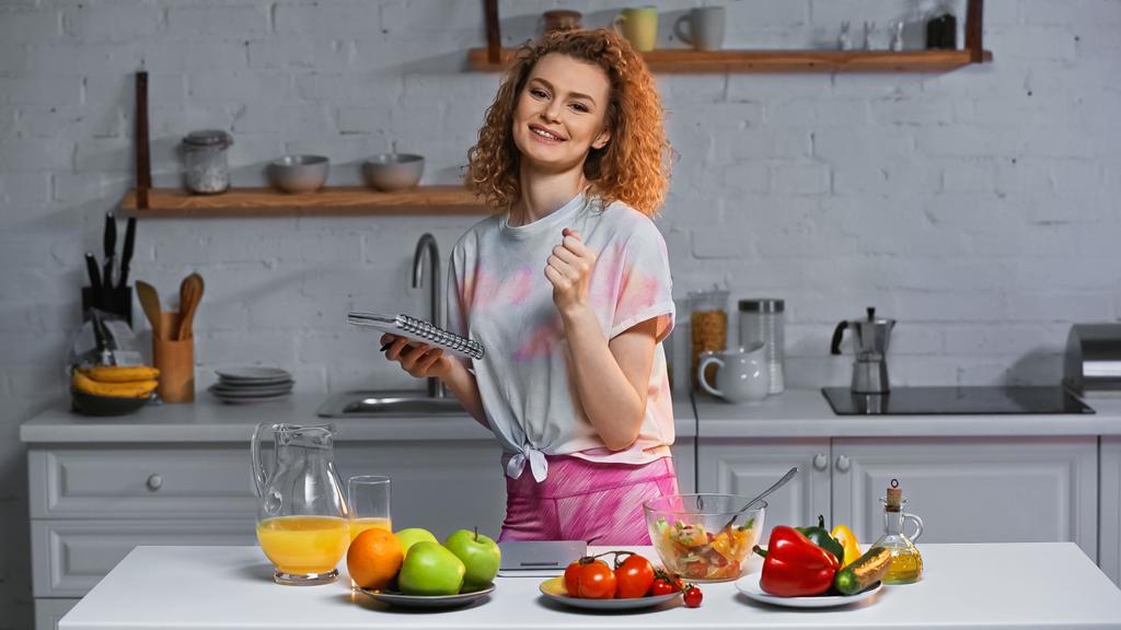 Улыбающаяся женщина держит ноутбук рядом с овощами и фруктами на кухонном столе  - Фото, изображение