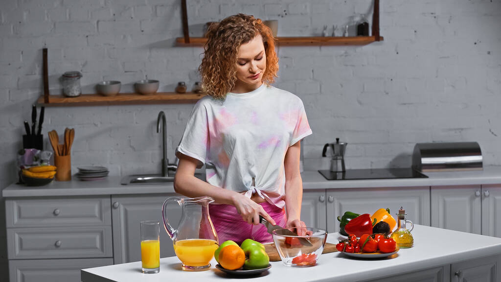 кудрявая молодая женщина режет свежие помидоры на кухне  - Фото, изображение