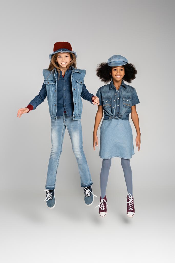 full length view of joyful multicultural girls in trendy denim clothes levitating on grey - Photo, Image