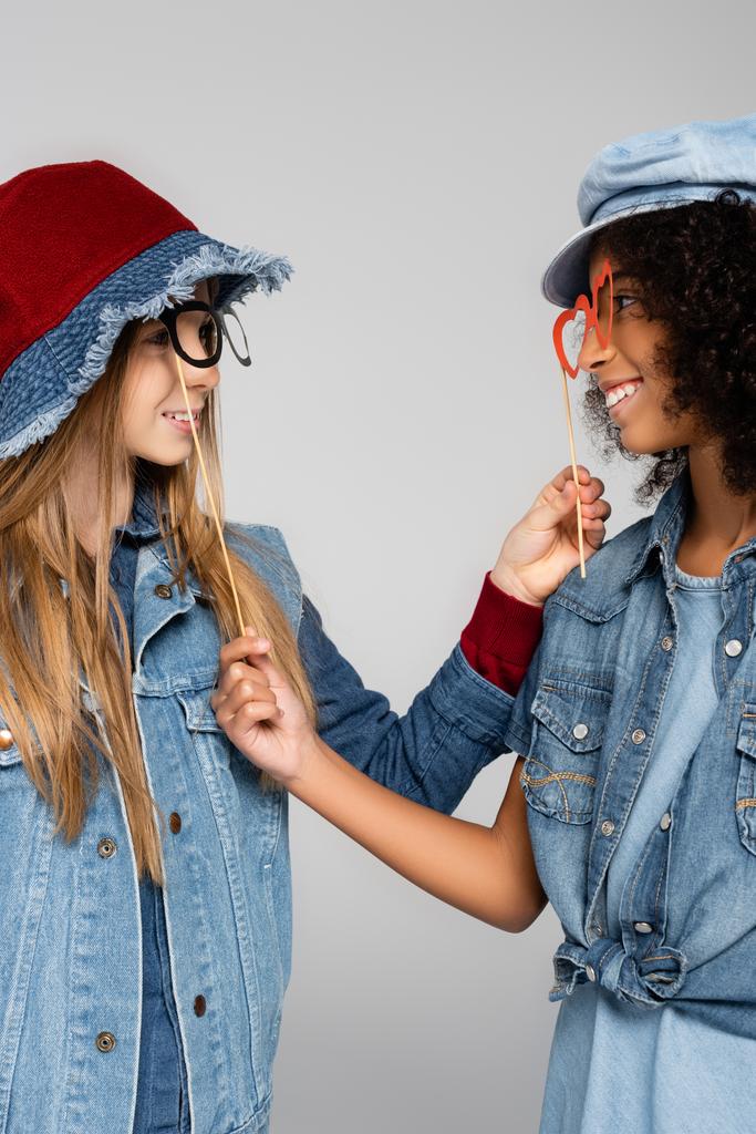 amigos multiculturales de moda con gafas de fiesta sonriendo el uno al otro aislados en gris - Foto, imagen