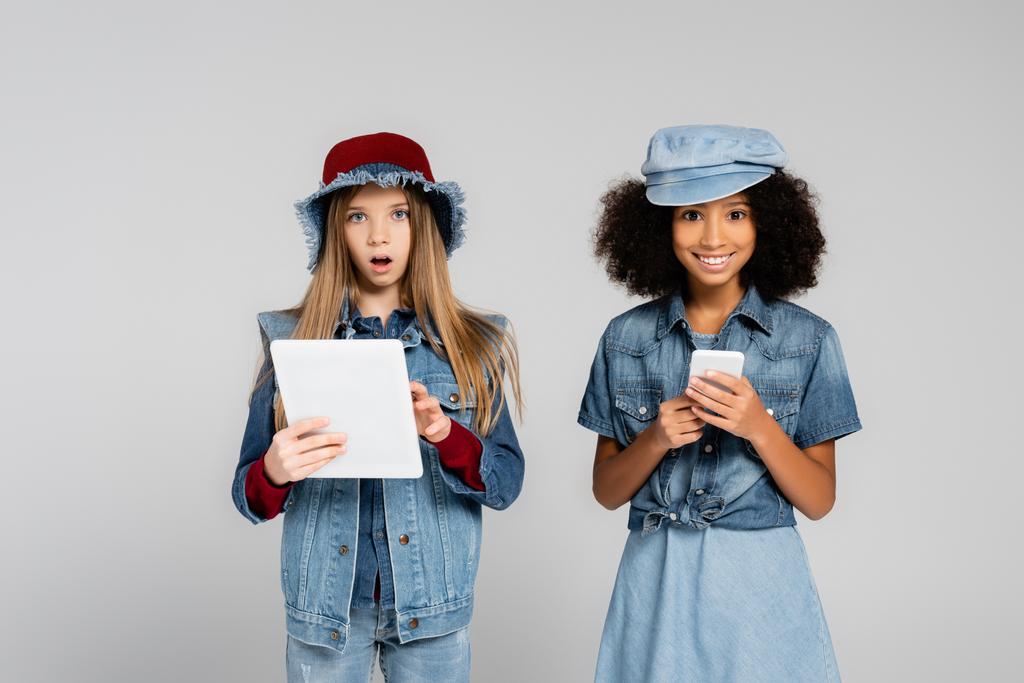 excited interracial kids in trendy clothes using smartphone and digital tablet isolated on grey - Photo, Image
