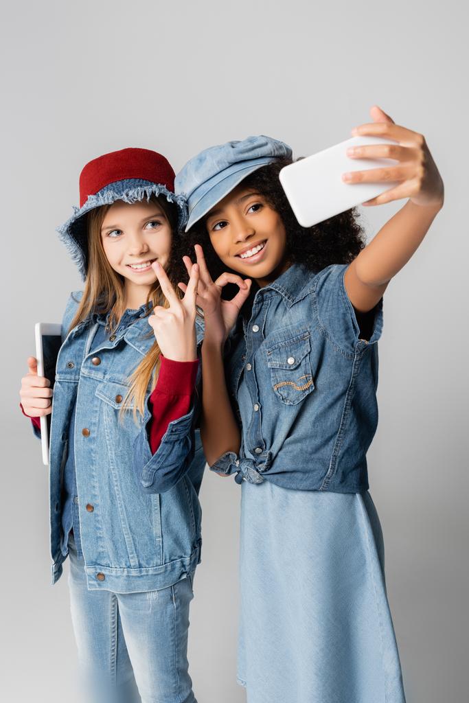fashionable interracial girls showing okay and victory signs while taking selfie on grey - Photo, Image