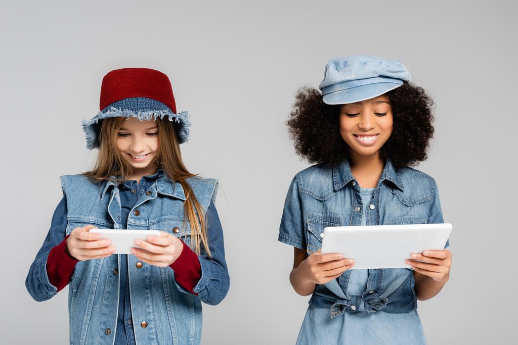 χαρούμενα διαφυλετικά παιδιά με μοντέρνα denim ρούχα και καπέλα χρησιμοποιώντας smartphone και ψηφιακό tablet που απομονώνονται σε γκρι - Φωτογραφία, εικόνα