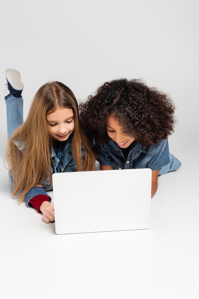 vrolijk interraciale vrienden in denim kleding met behulp van laptop terwijl liggend op grijs - Foto, afbeelding