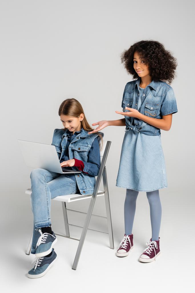 улыбающаяся, модная африканская американская девушка, указывающая на друга, сидящего на стуле и использующего ноутбук на сером - Фото, изображение