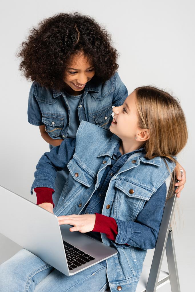 vrolijk Afrikaans amerikaans meisje in denim kleding in de buurt gelukkig vriend zitten op stoel met laptop geïsoleerd op grijs - Foto, afbeelding