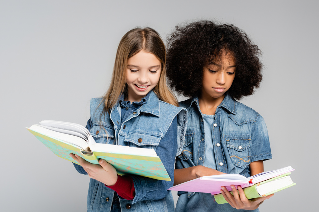 twee interraciale schoolmeisjes in modieuze denim kleding lezen boeken geïsoleerd op grijs - Foto, afbeelding