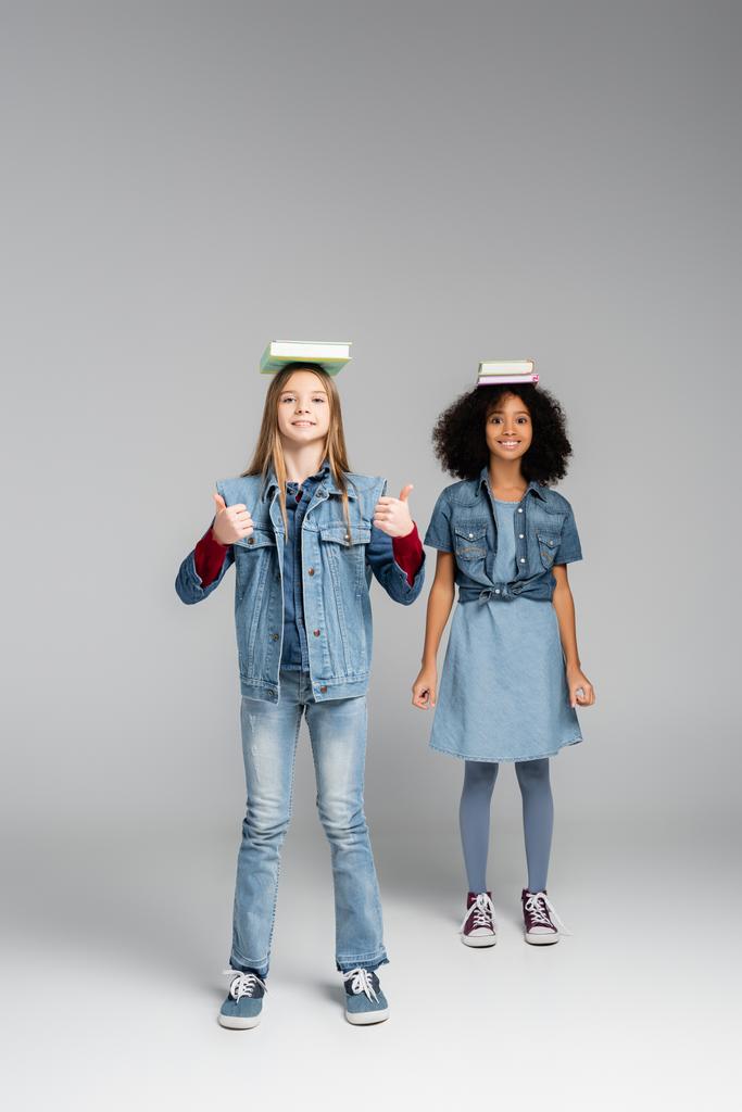 smiling interracial schoolgirls in denim clothes and gumshoes standing with books on heads on grey - Photo, Image