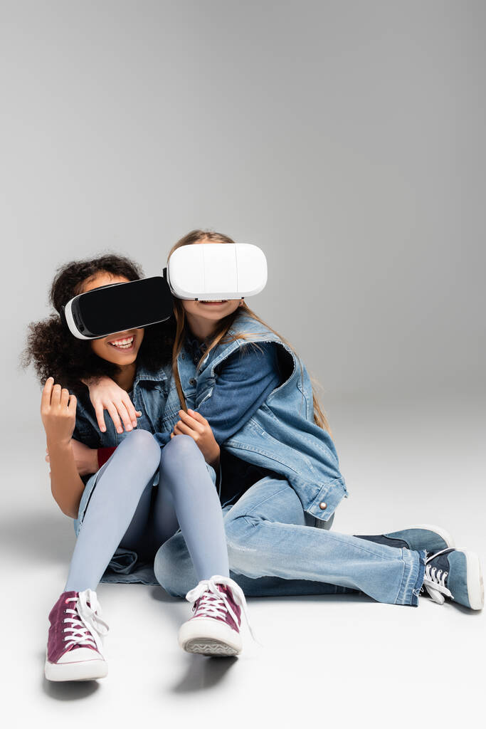 verängstigte und begeisterte multikulturelle Kinder in vr Headsets, die sich umarmen, während sie auf grau sitzen - Foto, Bild