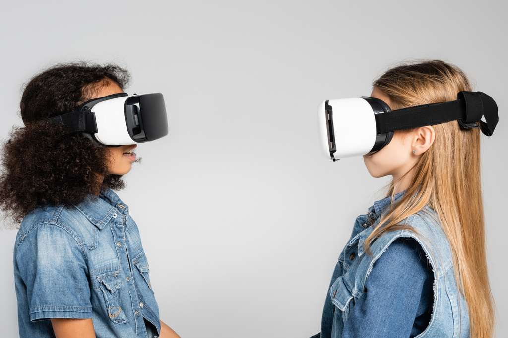 боковой вид модных межрасовых детей в гарнитурах VR, смотрящих друг на друга изолированно на сером фоне - Фото, изображение