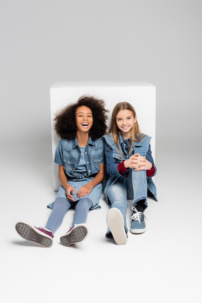 схвильовані міжрасові діти в джинсовому одязі, що сидить біля білої коробки на сірому
 - Фото, зображення