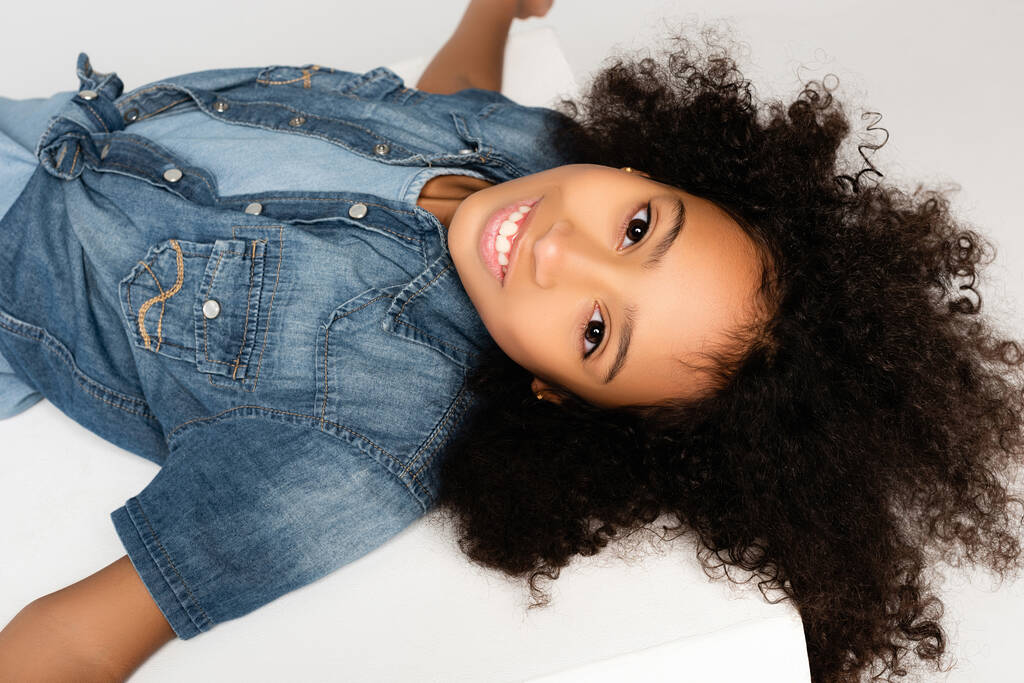 heureux afro-américaine fille en jean élégant vêtements souriant à la caméra tout en étant couché sur la surface blanche isolé sur gris - Photo, image
